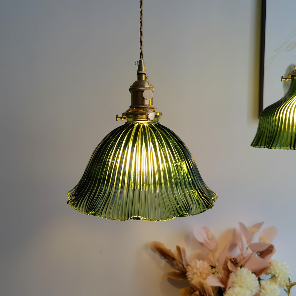 Lit green ribbed glass shade with a scalloped edge, hanging from a brass fitting beside a vase of pink dried flowers, from Clérachic.