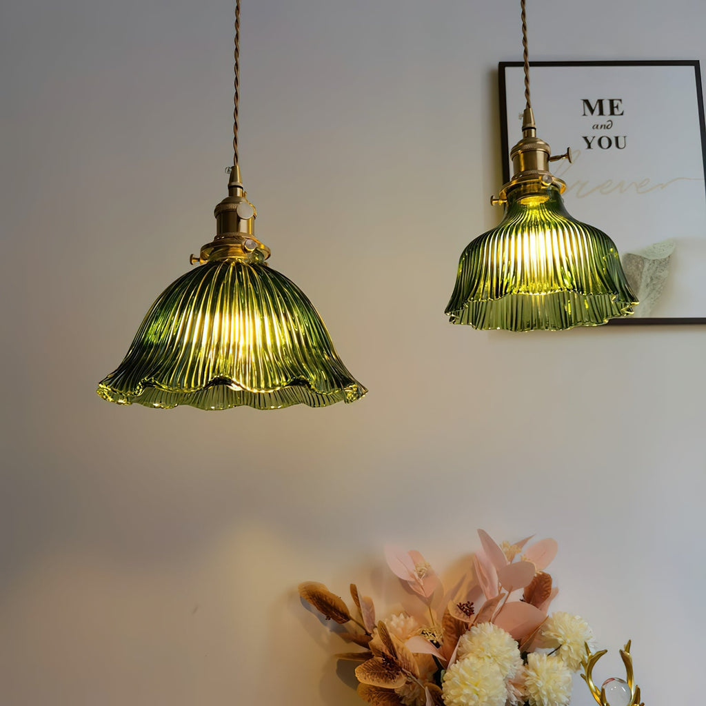 Two Clérachic green glass pendants lit side by side against a pale wall, a framed print and pink flowers behind them.