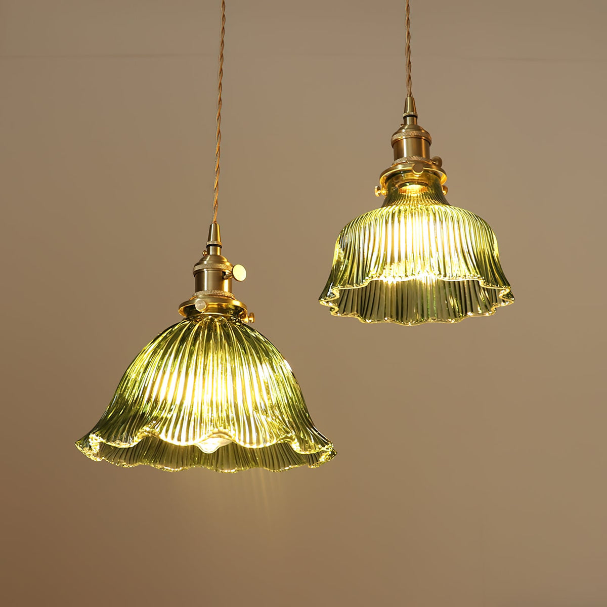 Two green glass pendants in different sizes, lit warm against a soft beige wall, both from Clérachic with brass fittings.