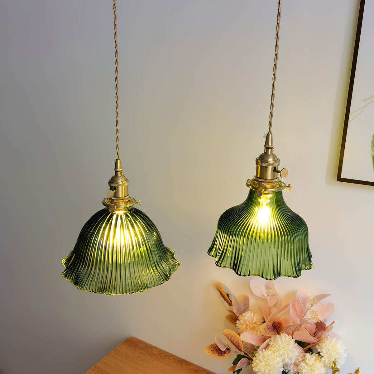 Pair of Clérachic green ribbed pendants lit above a wooden table, with pink flowers and a framed picture to one side.