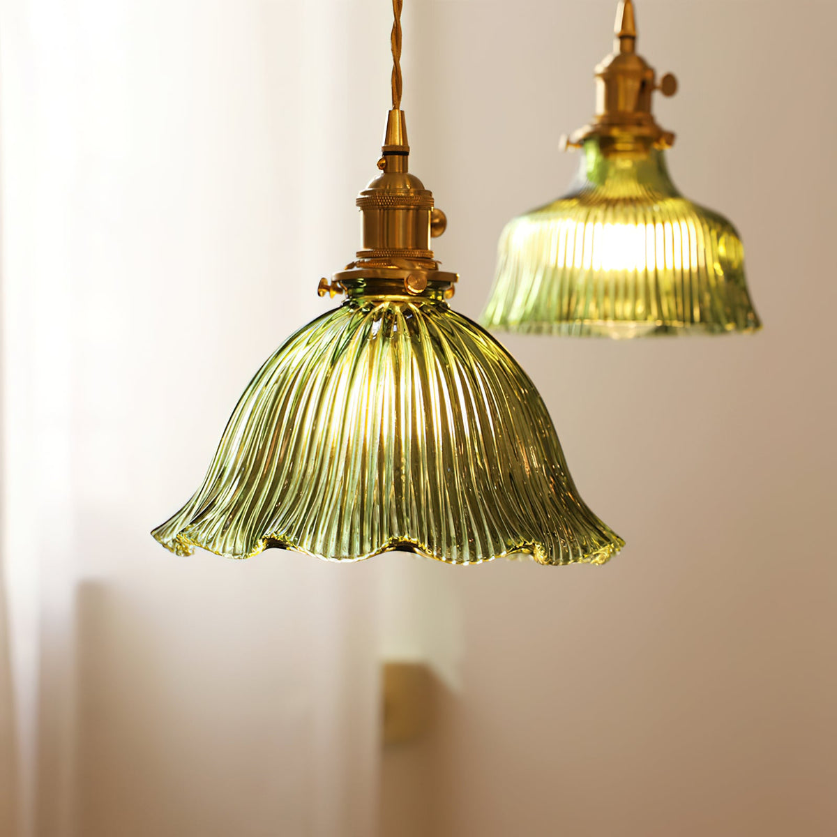 Two green glass pendants in different sizes, glowing against a cream wall beside a sheer curtain, both from Clérachic.