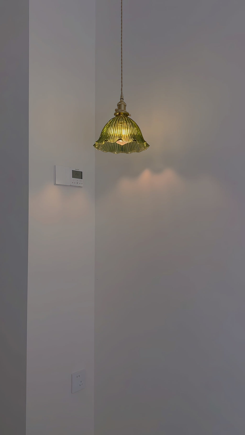 Distant view of a lit green glass pendant in a grey corner, casting soft light on the wall, from Clérachic.