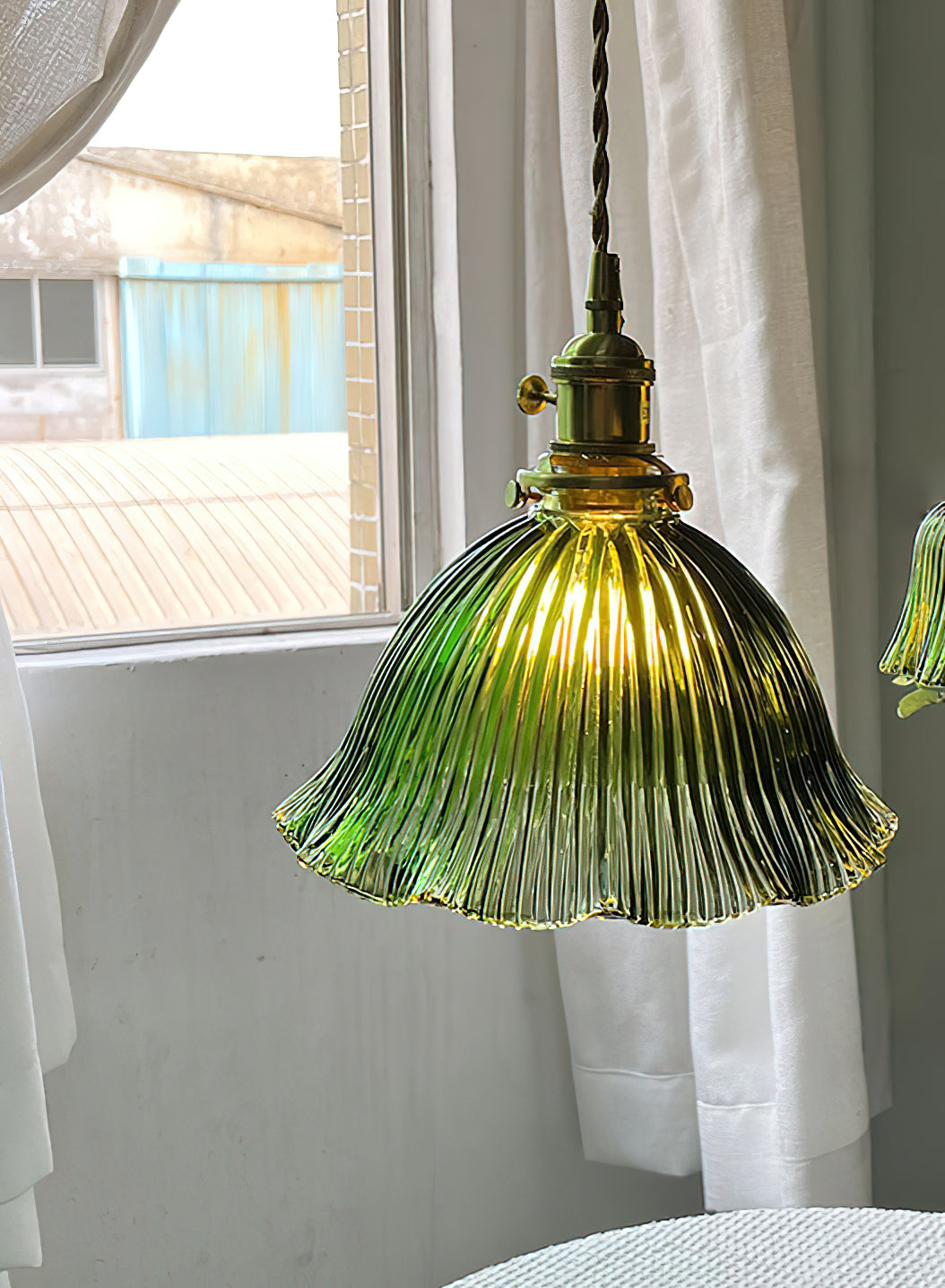 Lit green glass pendant by a bright window with white curtains, its ribbed shade glowing warm, from Clérachic.