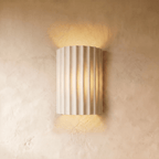 Clérachic wall lamp in white, its fluted half-cylinder shade glowing warm up and down a plaster wall.