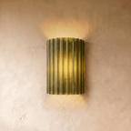 Clérachic wall lamp in olive green, fluted half-cylinder shade lit warm against a plain plaster wall.