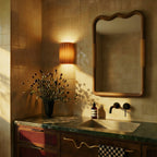 Clérachic wall lamp in burnt orange lit above a bathroom vanity with green marble and a wavy wood mirror.