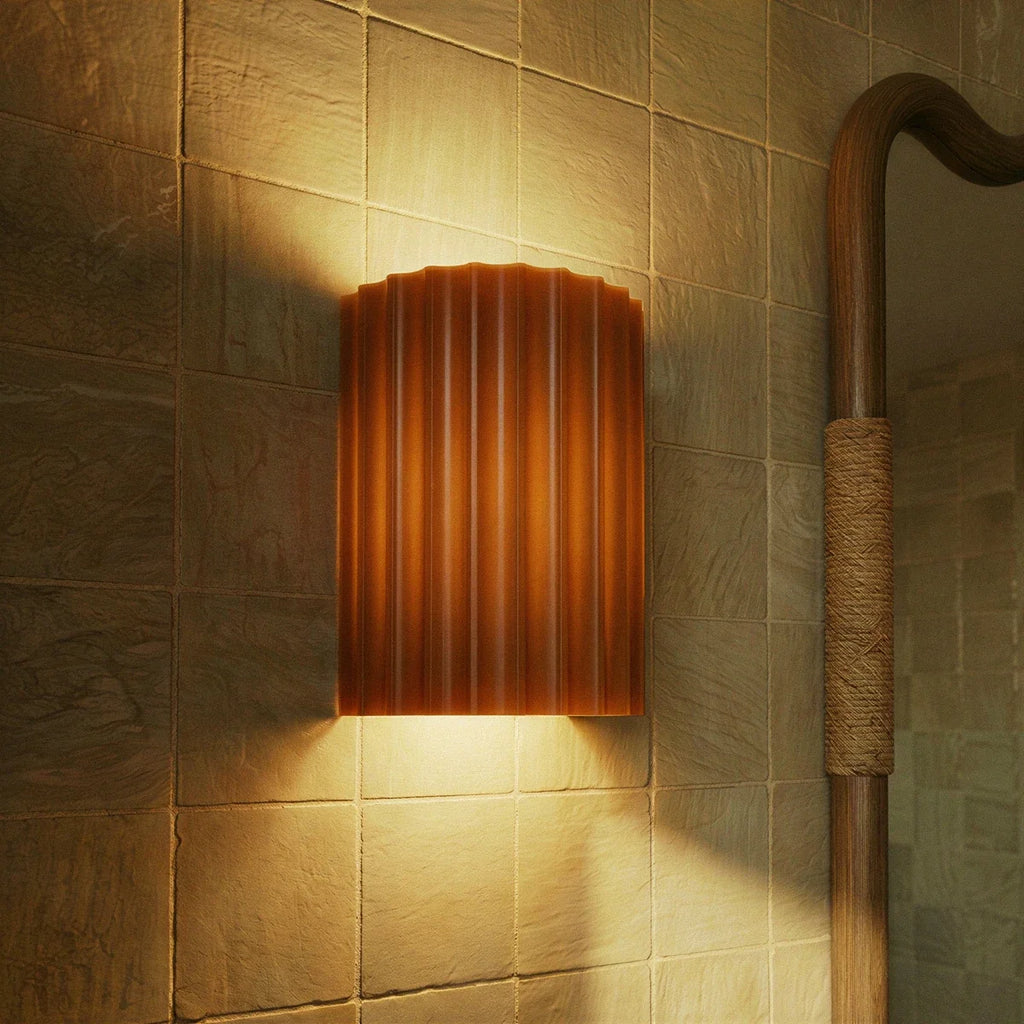 Clérachic wall lamp in burnt orange lit warm on ridged tile, beside a rope-wrapped mirror frame in a bathroom.