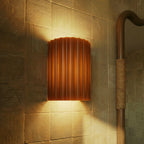 Clérachic wall lamp in burnt orange lit warm on ridged tile, beside a rope-wrapped mirror frame in a bathroom.