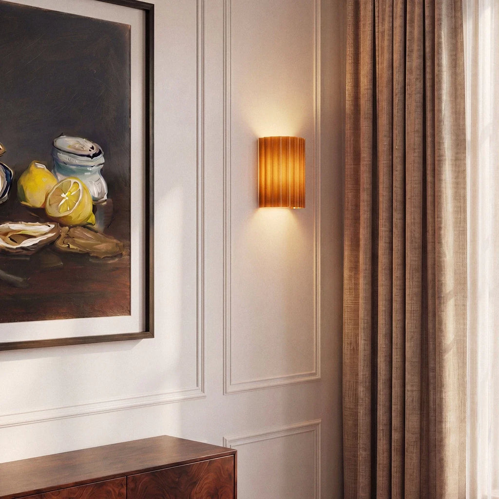 Clérachic wall lamp in burnt orange glowing beside a framed still-life painting and linen curtain.