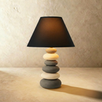 Clérachic grey ceramic table lamp with stacked pebble base and black fabric shade, glowing warm against a limewash wall.