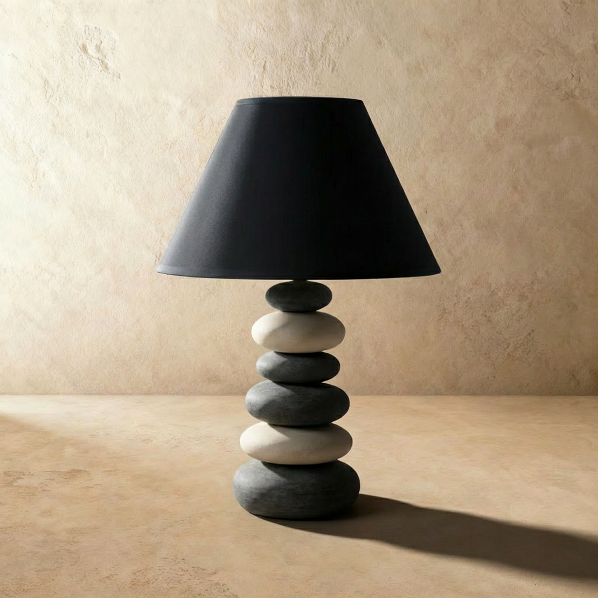 Clérachic table lamp unlit: six stacked grey and cream ceramic pebbles under a tapered black fabric shade on a sand surface.