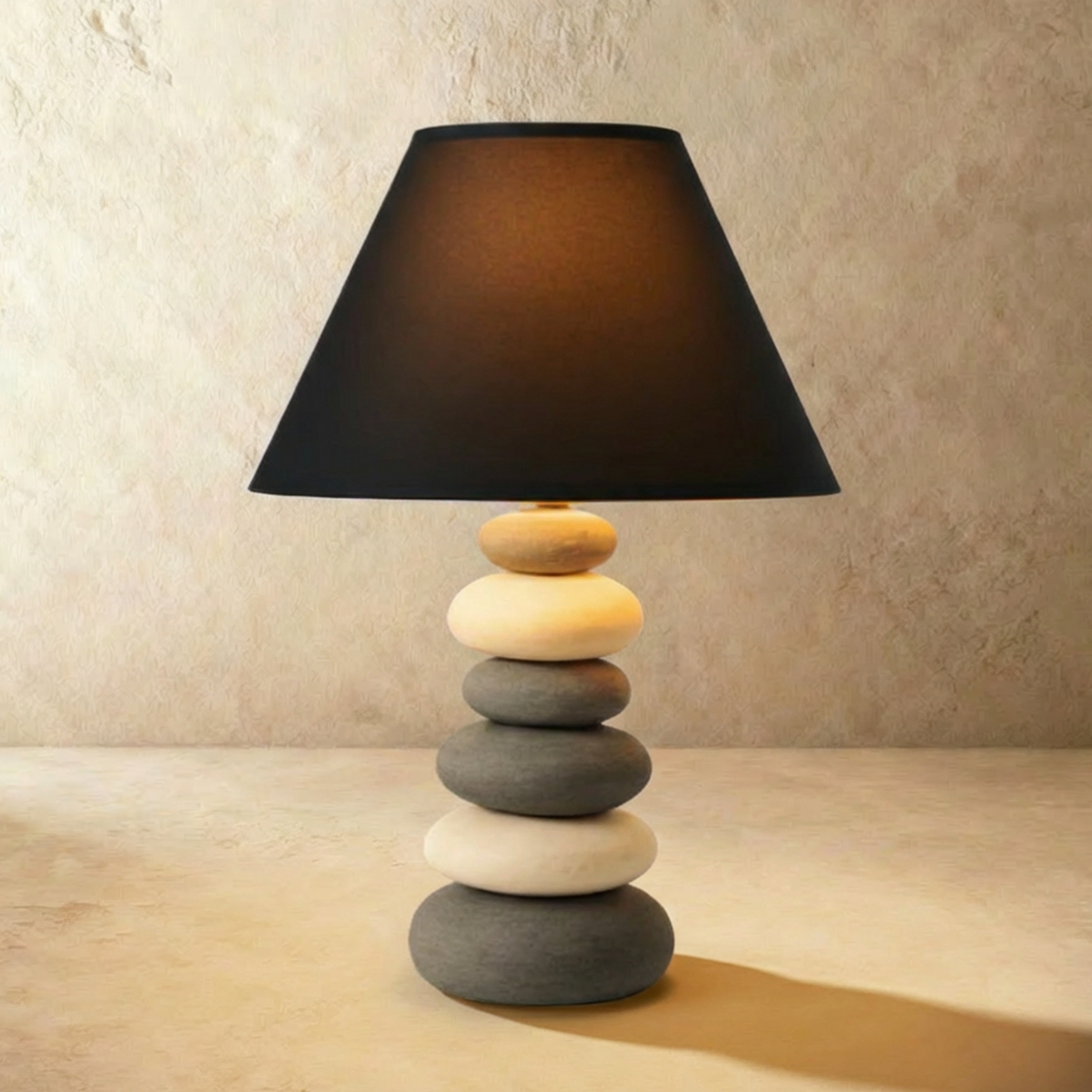 Clérachic larger pebble-stack ceramic table lamp with a wide black fabric shade, lit warm on a pale stone-effect backdrop.