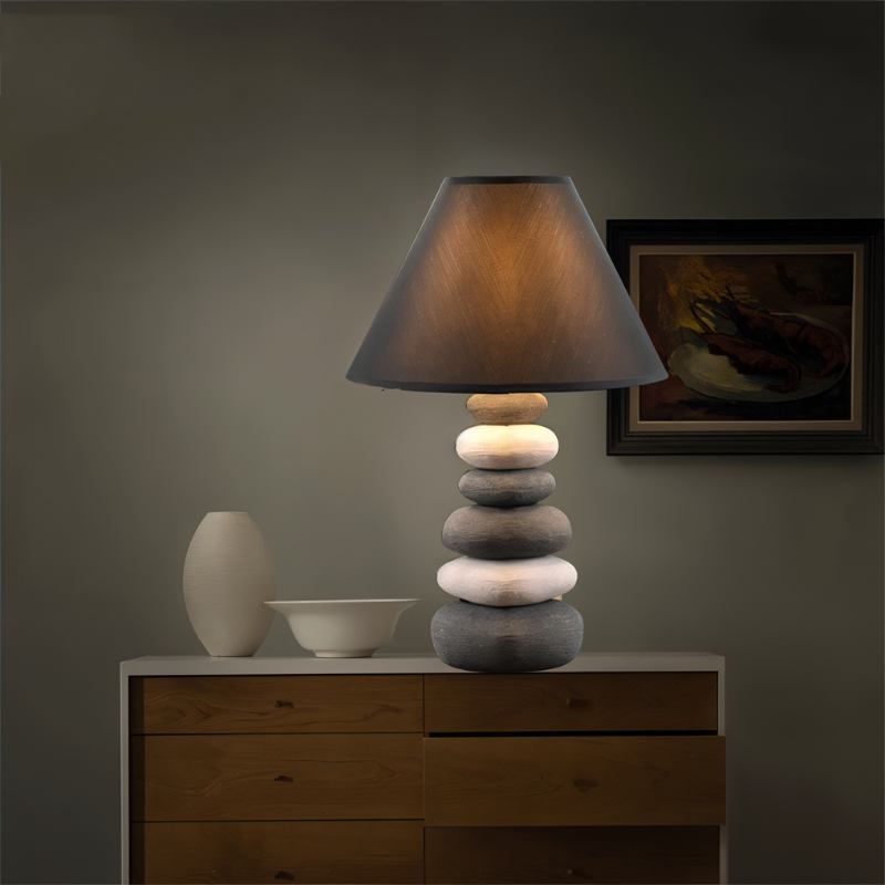 Clérachic pebble-stack ceramic table lamp lit on a wooden chest of drawers beside a white vase and bowl, framed art behind.