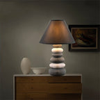 Clérachic pebble-stack ceramic table lamp lit on a wooden chest of drawers beside a white vase and bowl, framed art behind.