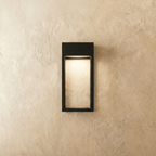 Clérachic grey steel wall sconce lit from within, warm light filling its open rectangular frame on a plaster wall.