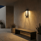 Clérachic grey steel wall sconce glowing warmly above a wooden bench and potted plants on an evening patio.