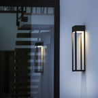 Taller Clérachic grey steel wall sconce glowing beside a glass entry door, reflecting an interior staircase at night.