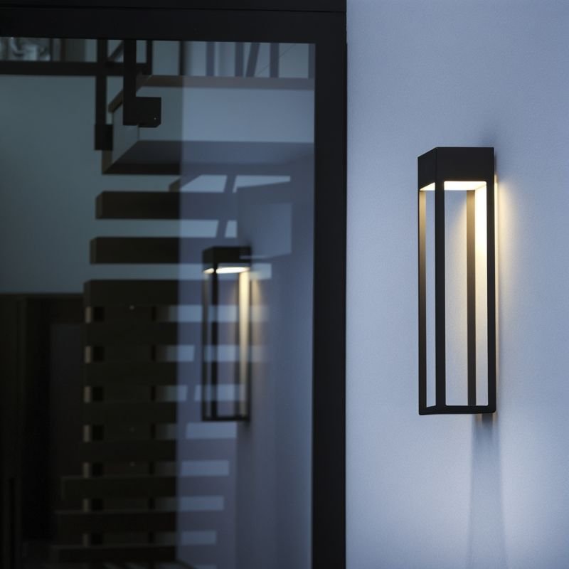 Taller Clérachic grey steel wall sconce glowing beside a glass entry door, reflecting an interior staircase at night.