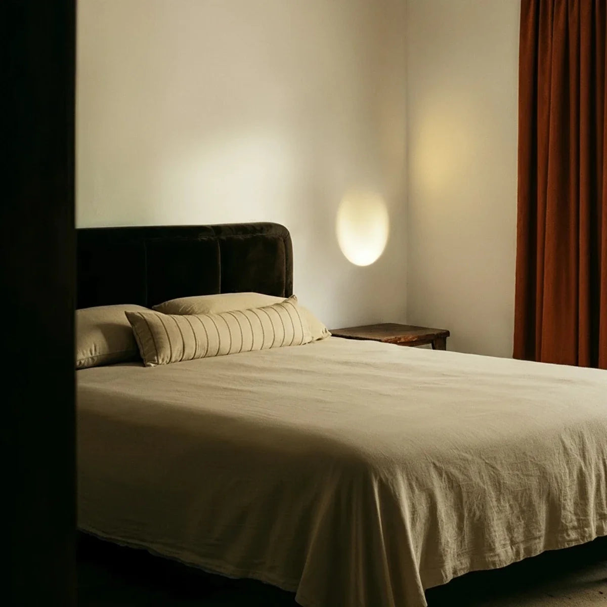 A Clérachic trimless wall sconce glows softly in a bedroom corner beside a dark velvet headboard at dusk.