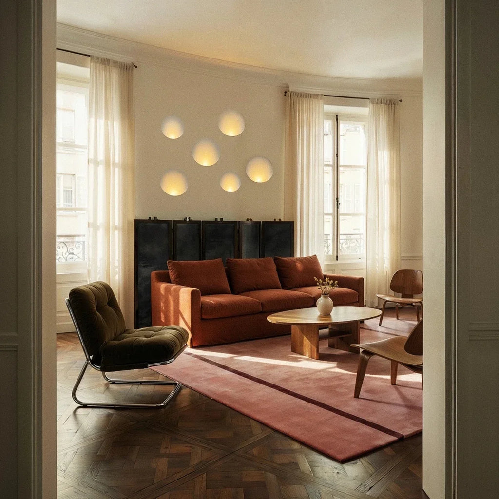 Six Clérachic trimless wall sconces form a cluster of glowing ovals above a rust-red sofa in a sunlit room.