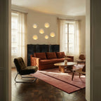 Six Clérachic trimless wall sconces form a cluster of glowing ovals above a rust-red sofa in a sunlit room.