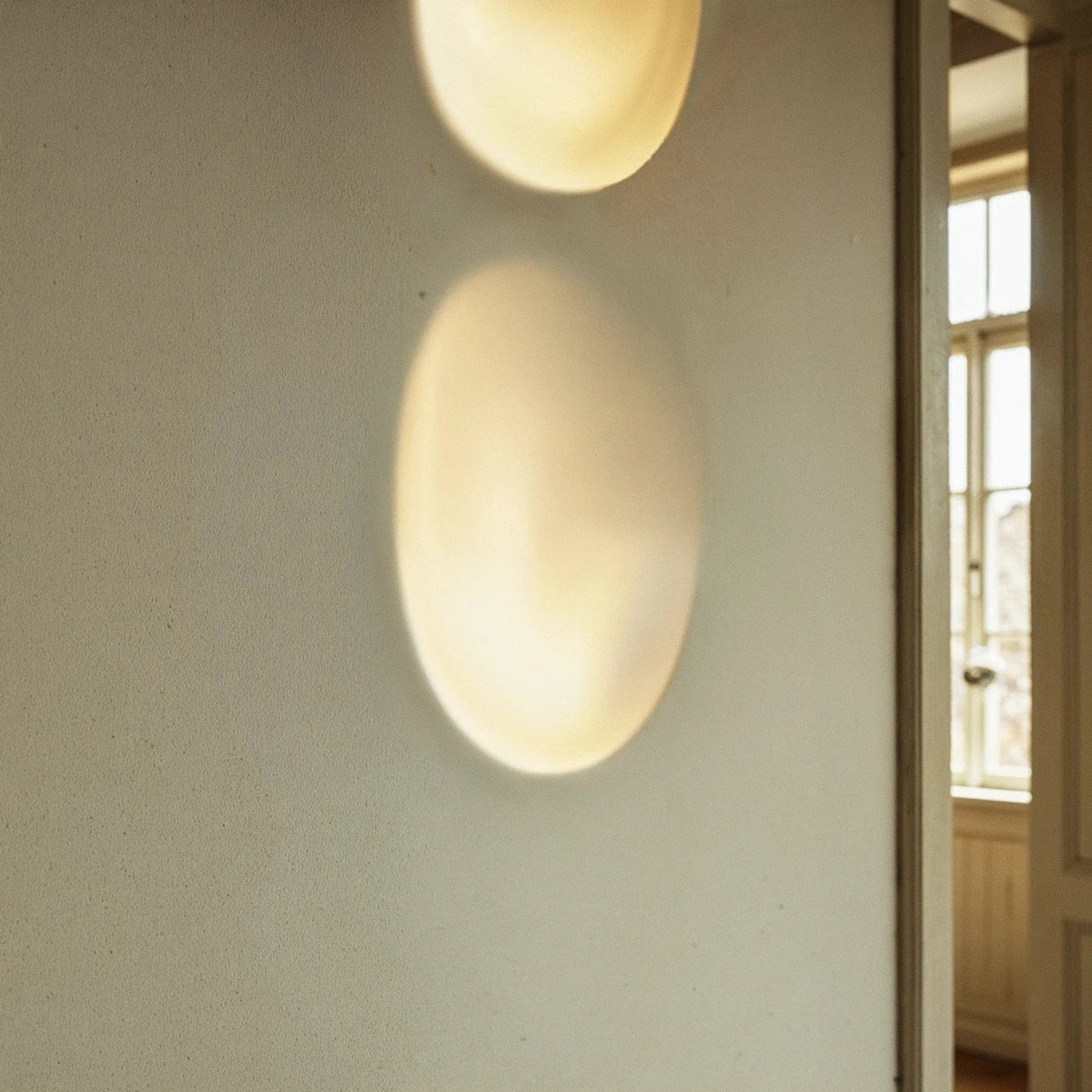This tight crop highlights the Clérachic sconce's warm glow stacked low on a plaster wall beside a window.