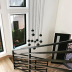 Clérachic matt black glass-globe pendants hung low beside a black railing in a stairwell with tall windows.