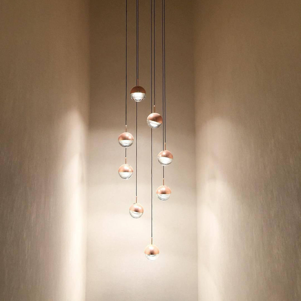Clérachic pendants with rose gold caps and clear glass lower halves hung at stepped heights down a warm-lit, tall stairwell wall.