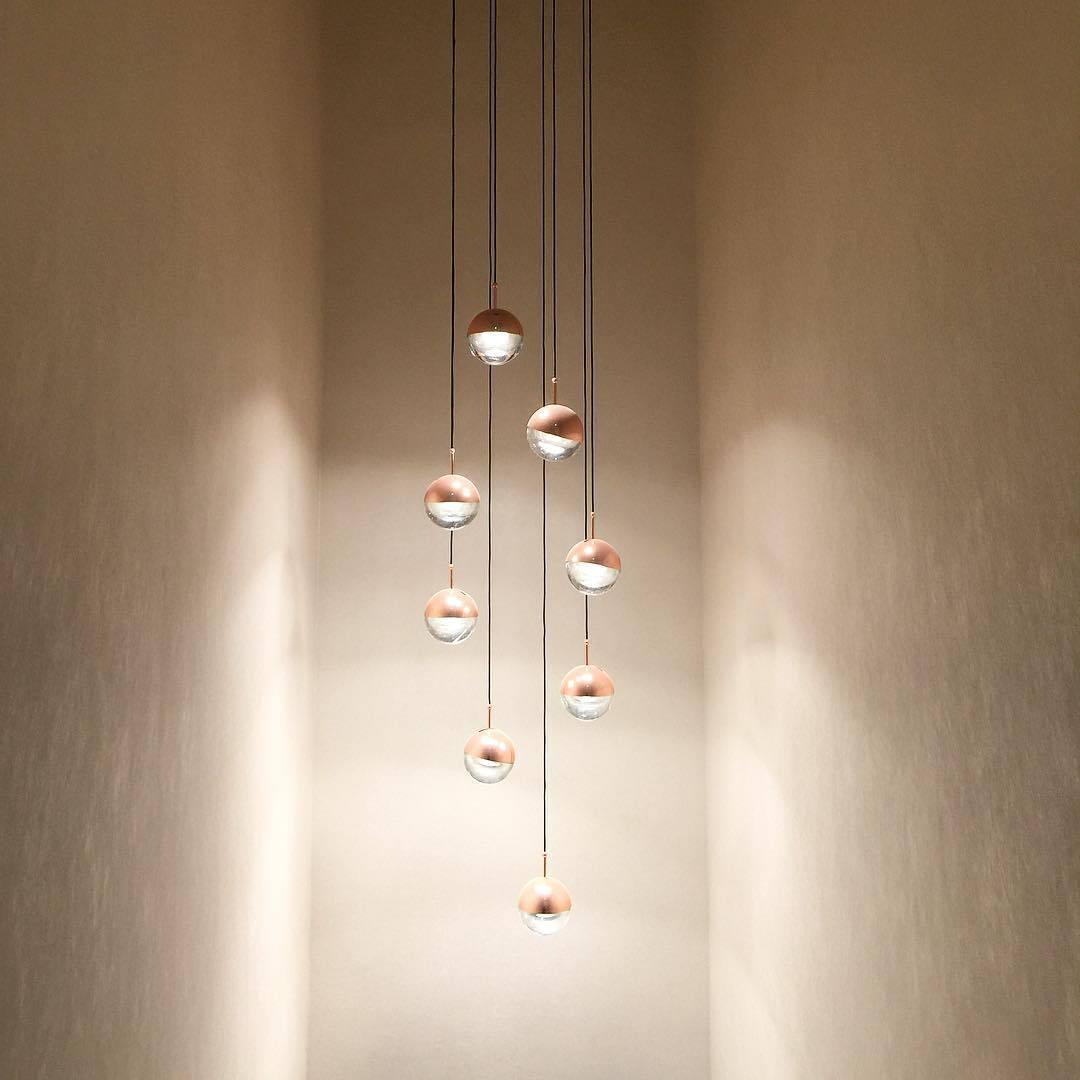 Clérachic pendants with rose gold caps and clear glass lower halves hung at stepped heights down a warm-lit, tall stairwell wall.