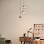 Three rose gold Clérachic globe pendants at different heights above a wooden console, a framed print and a vintage camera.