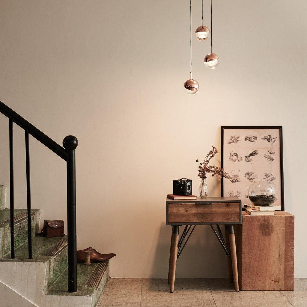 Three rose gold Clérachic globe pendants above a rustic wooden console beside a staircase with a black banister.