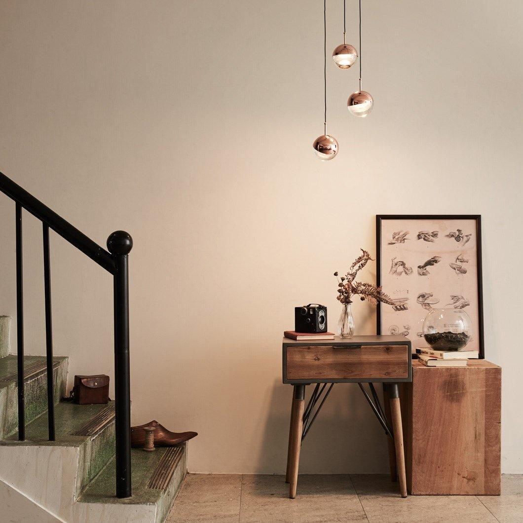 Three rose gold Clérachic globe pendants above a rustic wooden console beside a staircase with a black banister.