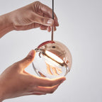 Hands holding a rose gold Clérachic globe pendant, its clear glass half lit warmly from inside the metal cap.