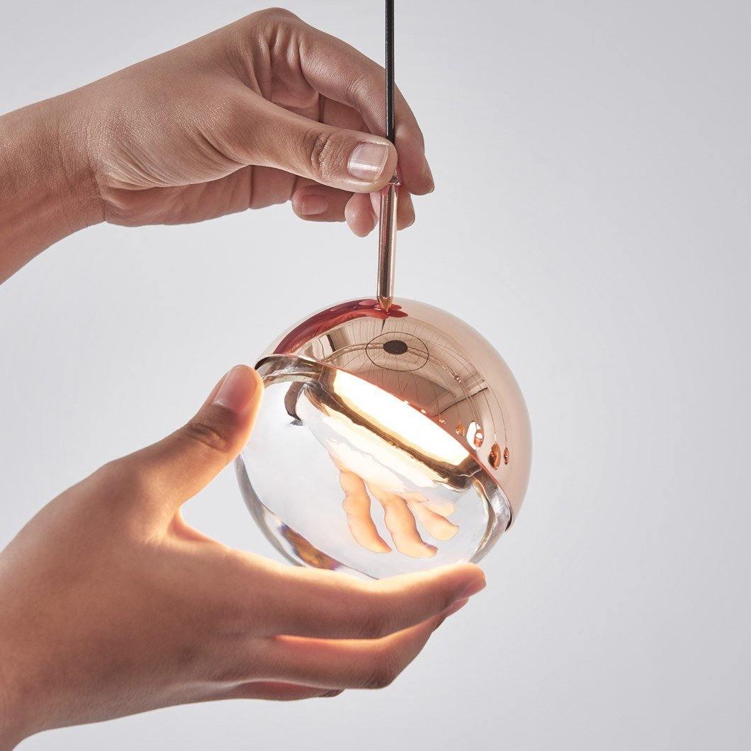 Hands holding a rose gold Clérachic globe pendant, its clear glass half lit warmly from inside the metal cap.