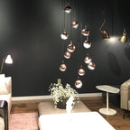 Many rose gold and clear Clérachic globe pendants glowing against a dark wall, above a white coffee table and a lamp.