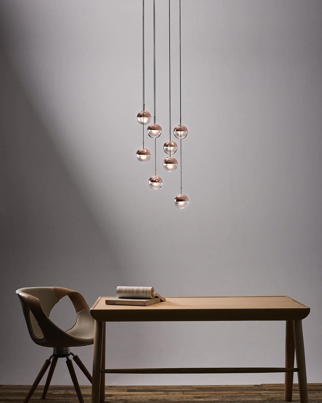 Cluster of rose gold Clérachic globe pendants hung above a wooden desk and an upholstered chair, against a grey wall.