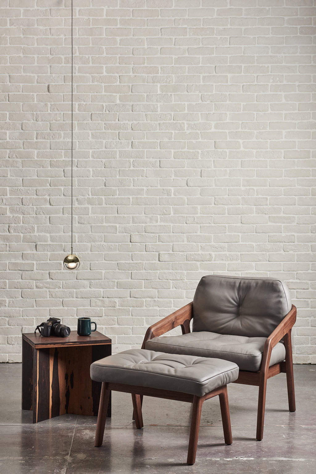 Single gold-finish Clérachic globe pendant against a white brick wall, above a leather armchair and a footstool.
