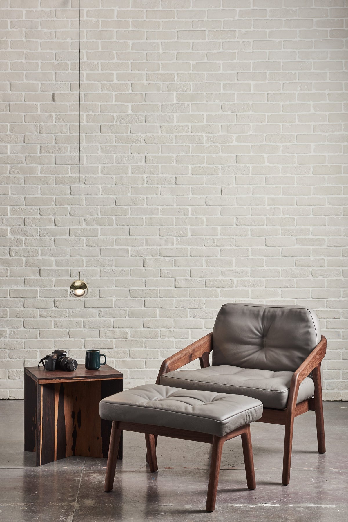 Single gold-finish Clérachic globe pendant against a white brick wall, above a leather armchair and a footstool.