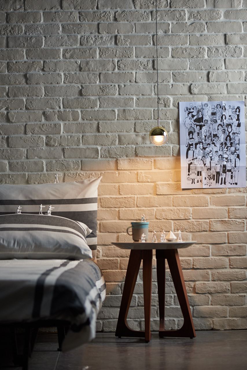 Gold-finish Clérachic globe pendant lighting an exposed brick wall, above a bed with a grey striped cushion.