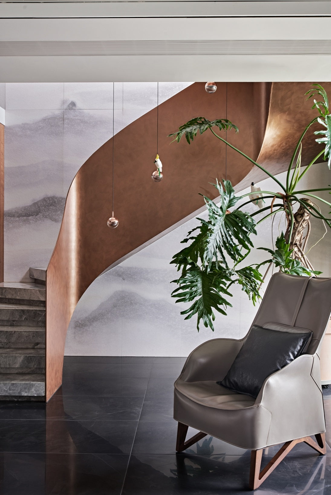 Small Clérachic globe pendants glimpsed beside a curved copper-toned staircase, above a leather chair and a large leafy plant.