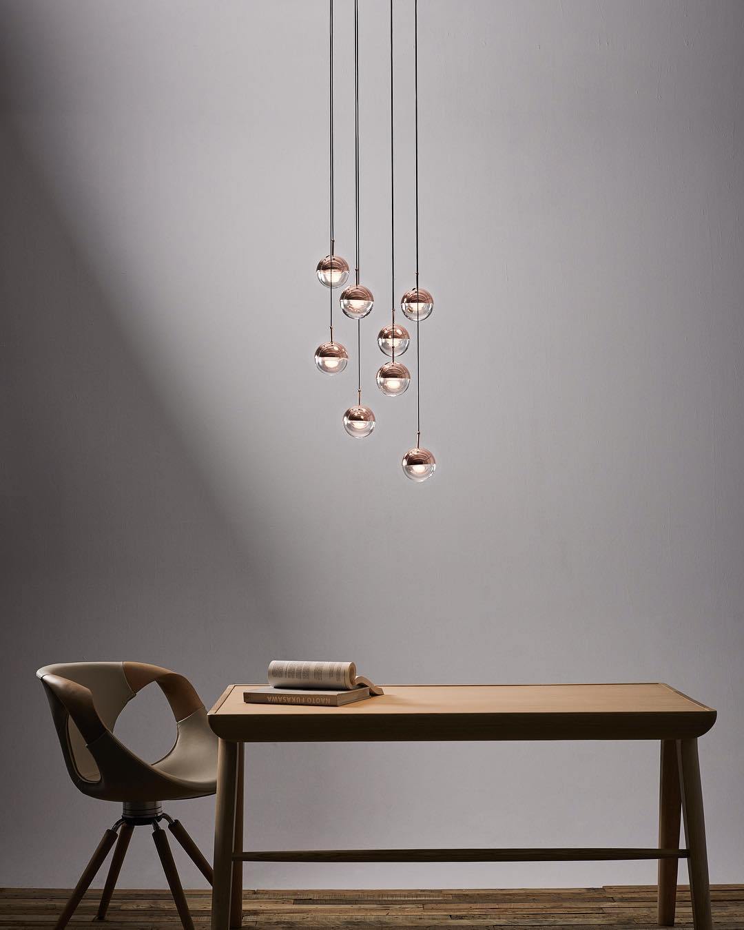 Cluster of rose gold Clérachic globe pendants against a grey wall, above a wooden desk and a moulded chair.
