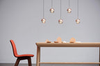 Five small rose gold Clérachic globe pendants hung in a loose group above a pale wooden table and a red chair.