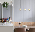 Three gold-finish Clérachic globe pendants in a row against a pale panelled wall, above a table with a wine glass.