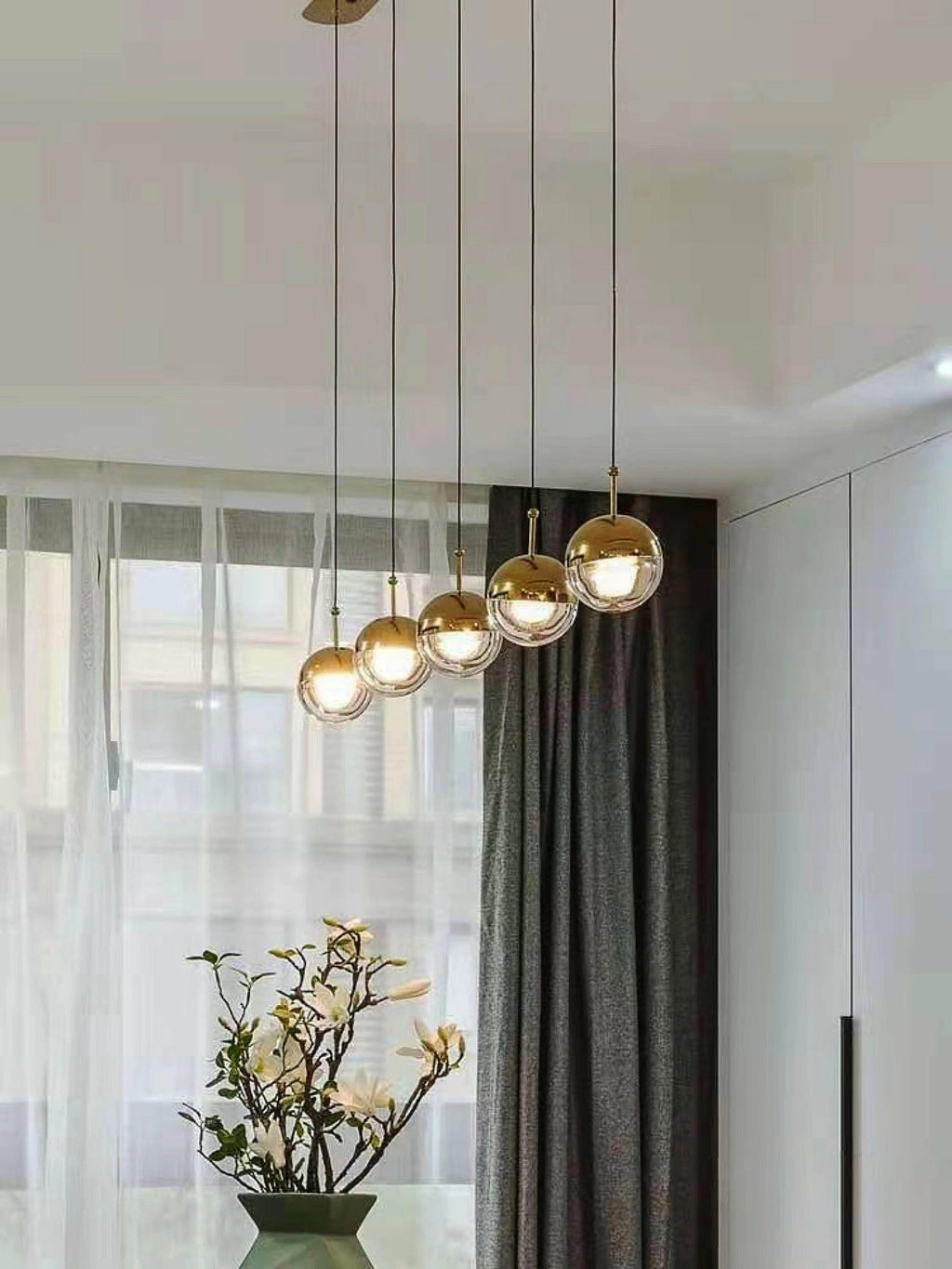 Row of five gold-finish Clérachic globe pendants rising on a diagonal beside a sheer curtain and a vase of white blossom.