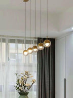 Row of five gold-finish Clérachic globe pendants rising on a diagonal beside a sheer curtain and a vase of white blossom.