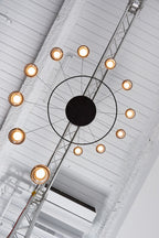 Looking up at a Clérachic ring chandelier with warm-lit globes around a black centre, beside a metal ceiling truss.