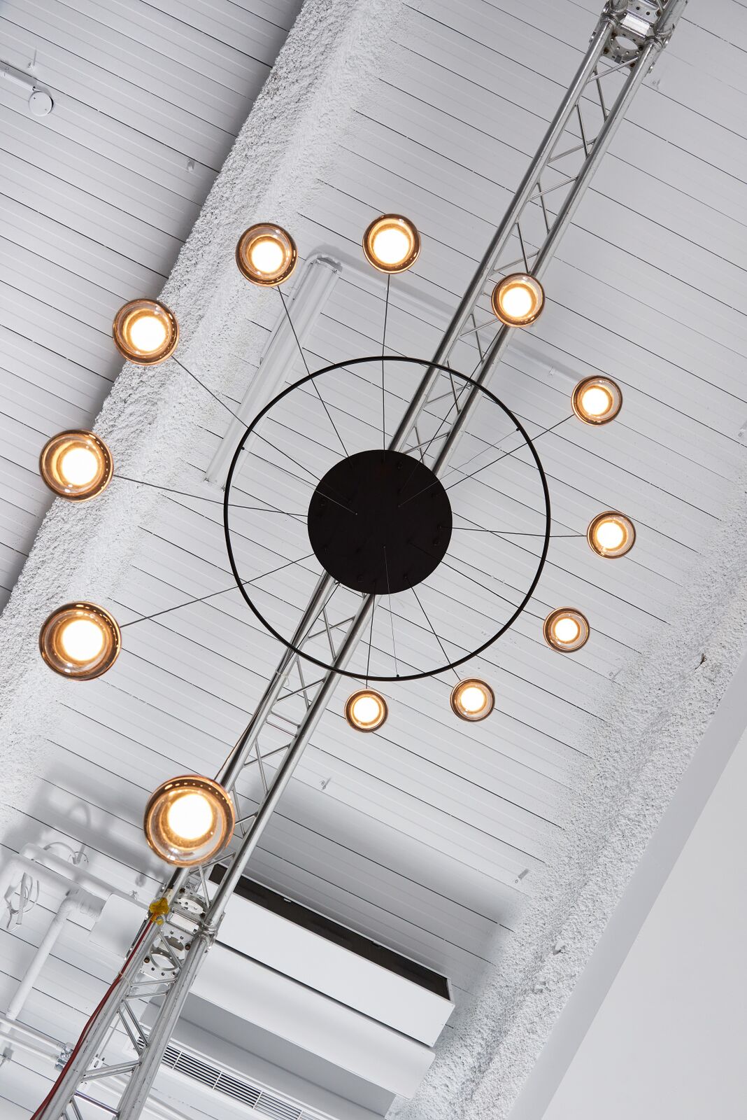 Looking up at a Clérachic ring chandelier with warm-lit globes around a black centre, beside a metal ceiling truss.