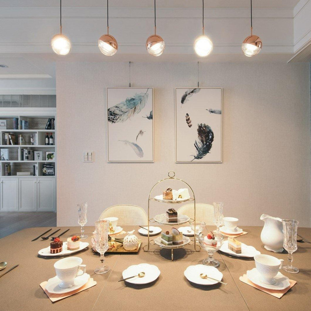 Row of five rose gold and clear Clérachic globe pendants above a laid afternoon-tea table with feather prints on the wall.