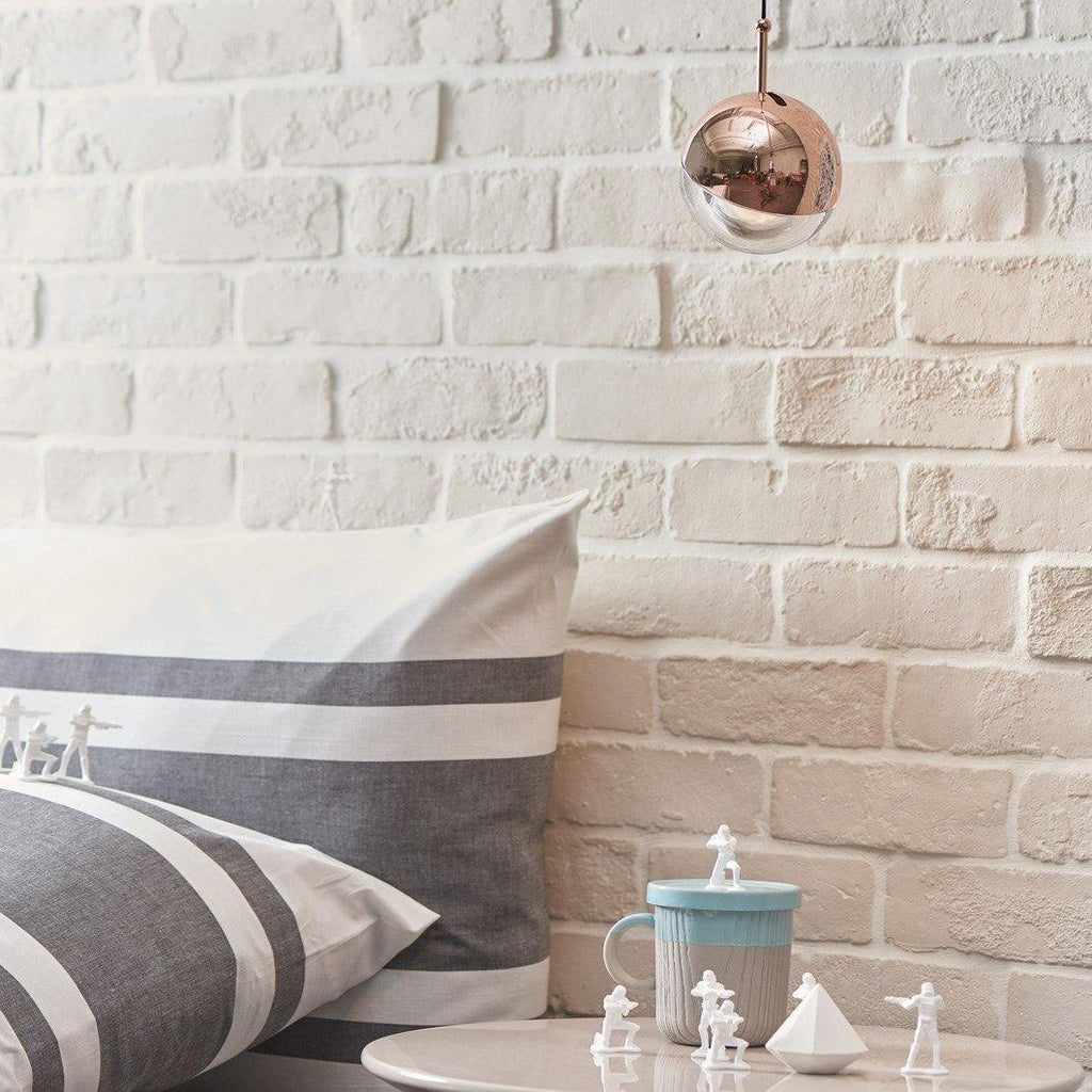 A rose gold mirror-finish Clérachic globe pendant against a white brick wall, above a striped cushion and small figurines.