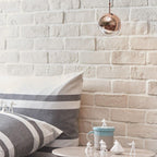 A rose gold mirror-finish Clérachic globe pendant against a white brick wall, above a striped cushion and small figurines.